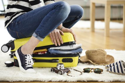 A young woman sitting on top of suitcase while trying to close it