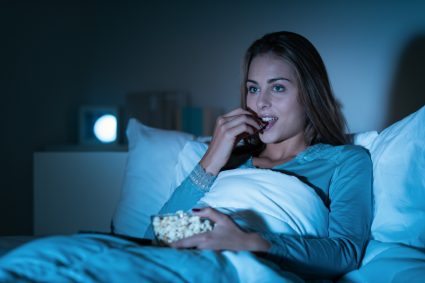 A woman sitting up in bed, eating popcorn and watching a movie