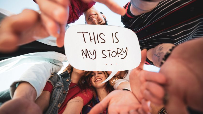 A diverse group of smiling students looking down into the camera, holding a speech bubble sign that says 'THIS IS MY STORY' at a gathering, symbolizing individuality and shared experiences in crafting personal narratives.